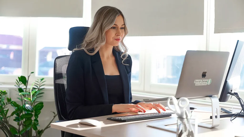 Blond kvinna sitter och arbetar framför en dator i en ljus kontorsmiljö.